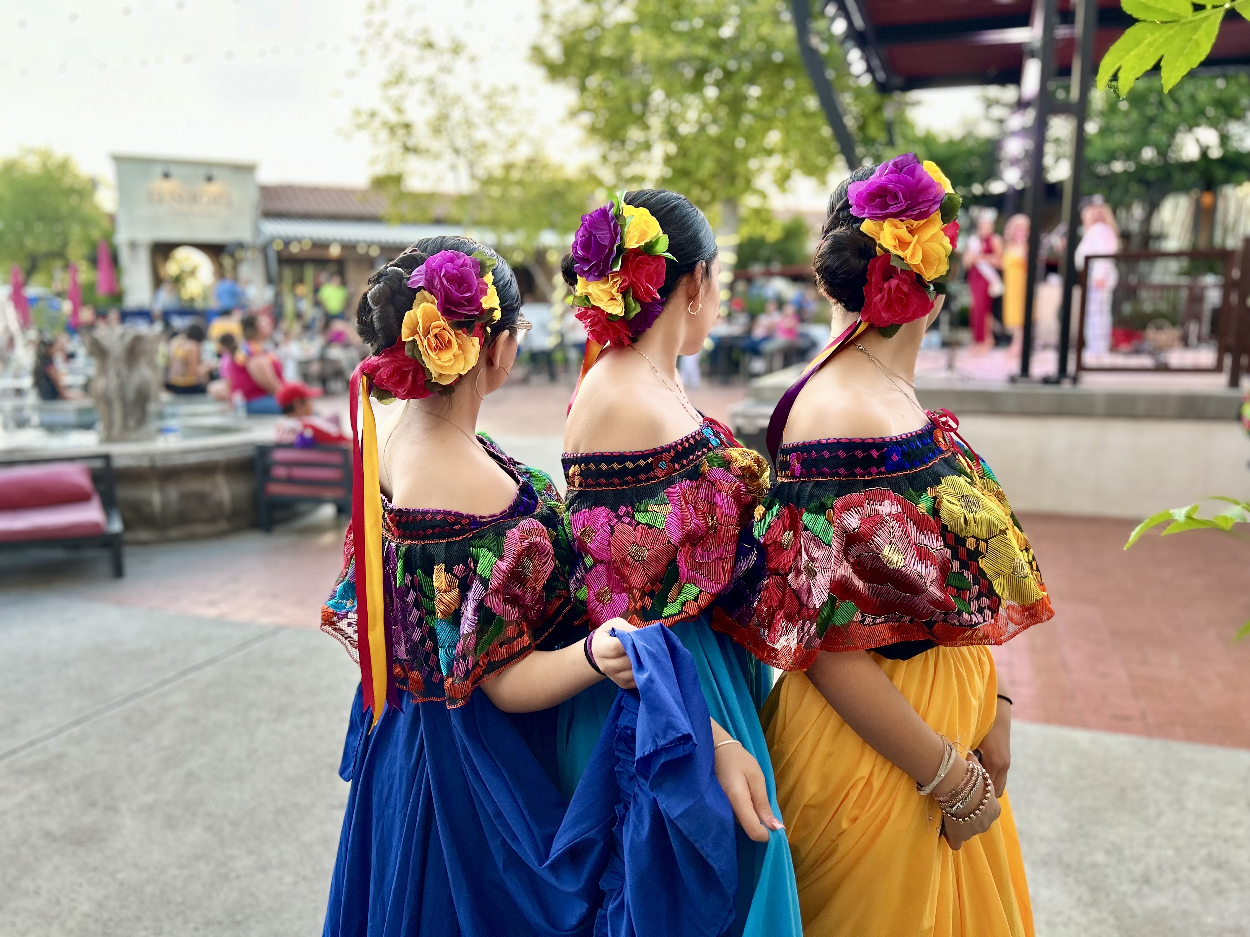 Three dark haired girls pose in their traditional colorful dresses and flower headpieces